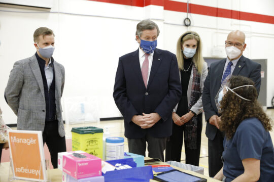 A group of 4 people standing up and wearing face masks talking to a nurse.
