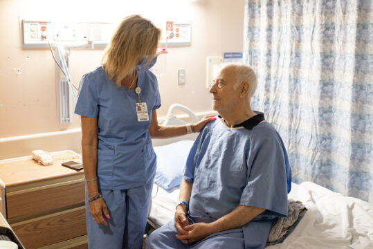 A health care worker is assisting an elderly patient who is sitting on a bed.