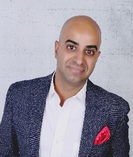 Board Member Aayaz Pira is photographed from the shoulders up in front of a grey background. He smiles at the camera. He has medium-tone skin. He wears a tweed dark grey suit, a white collared shirt and a red pocket square.