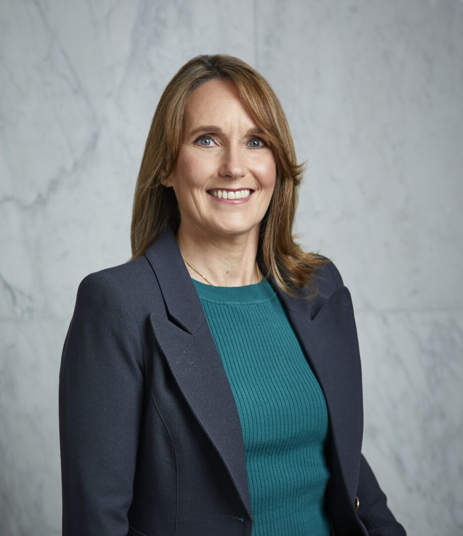 Board Chair, of the North York General Foundation, Aine O’Flynn is photographed from the waist down with arms at her side in front of a marbled grey background. She smiles at the camera. Her hair shoulder length hair is light brown. She has light-tone skin. She wears a dark grey suit with a teal sweater.