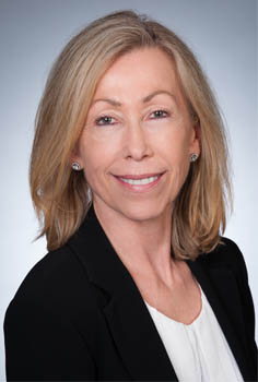 Board member Debbie Fisher is photographed from the shoulders up in front of a grey background. Se smiles at the camera. She has light-tone skin and blonde shoulder length hair. She wears a black suite with a white blouse.
