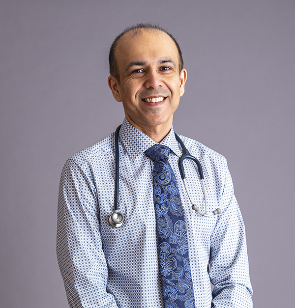 Dr. Ronik Kanani - Dr. Ronik Kanan is photographed from the waist up in front of a grey background. He smiles at the camera. He has light-tone skin and very short black hair. He wears a light blue collared shirt with a darker blue patterned tie. He also wears a stethoscope.