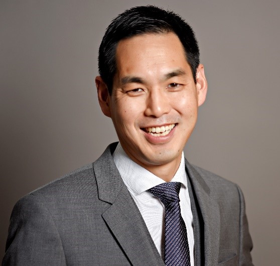 Phil Shin is photographed from the shoulders up in front of a grey background. He smiles at the camera. He has light-tone skin. He has short black hair. He wears a light grey suit with a white collared shirt and a dark blue tie.
