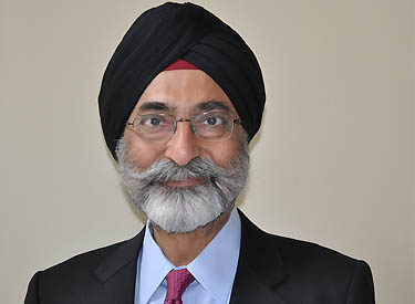 Harcharan (Harry) Singh is photographed from the shoulders up in front of a beige background. He smiles at the camera. He has medium-tone skin and a white bear. He wears a black suit with a light blue colored shirt and a red tie. He also wears glasses and a black turban.