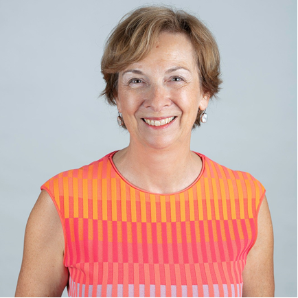 Board Member Janet Beed is photographer from the shoulders up in front of a grey background. She smiles at the camera. She has light-tone skin. She has short light brown hair and wears an orange and pink sleeveless top with horizonal stripes.
