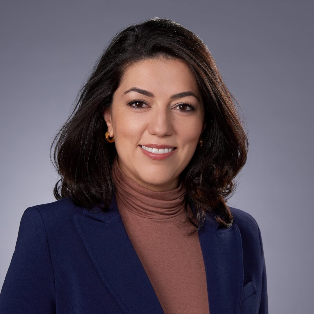 Dr. Maral Nadjafi - Dr. Maral Nadjafi is photographed from the shoulders up in front a grey background. She smiles at the camera. He has light-tone skin and dark brown shoulder length hair. She wears a black suit with a brown turtleneck.