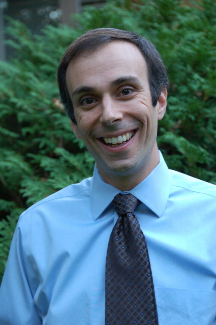 Board Member Dr. Benjamin Bell is photographed from the shoulders up outside in front of green foliage. He smiles at the camera. He has dark brown hair. He has light-tone skin. He wears a light blue collared shirt and a black tie.
