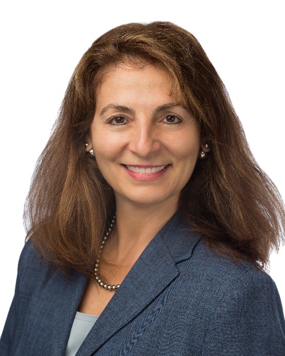 Board Member Toni Rossi is photographed from the shoulders up in front of white background. She smiles at the camera. She has light-tone skin. Se has shoulder length brown hair. She wears a dark blue suit with a light blue blouse.