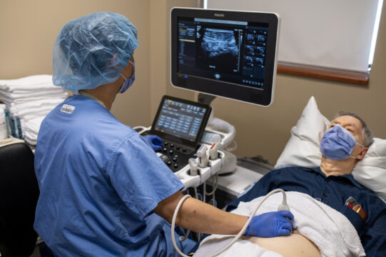 A health care professional using an ultrasound machine of a patient. The machine has data and medical imaging on display.