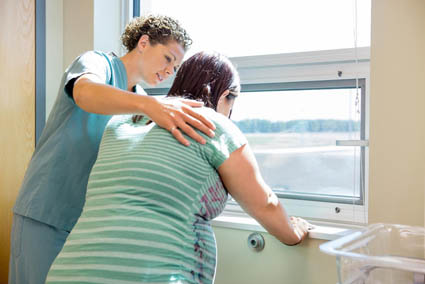 Mid adult female nurse comforting tensed pregnant woman leaning on window sill in hospital room