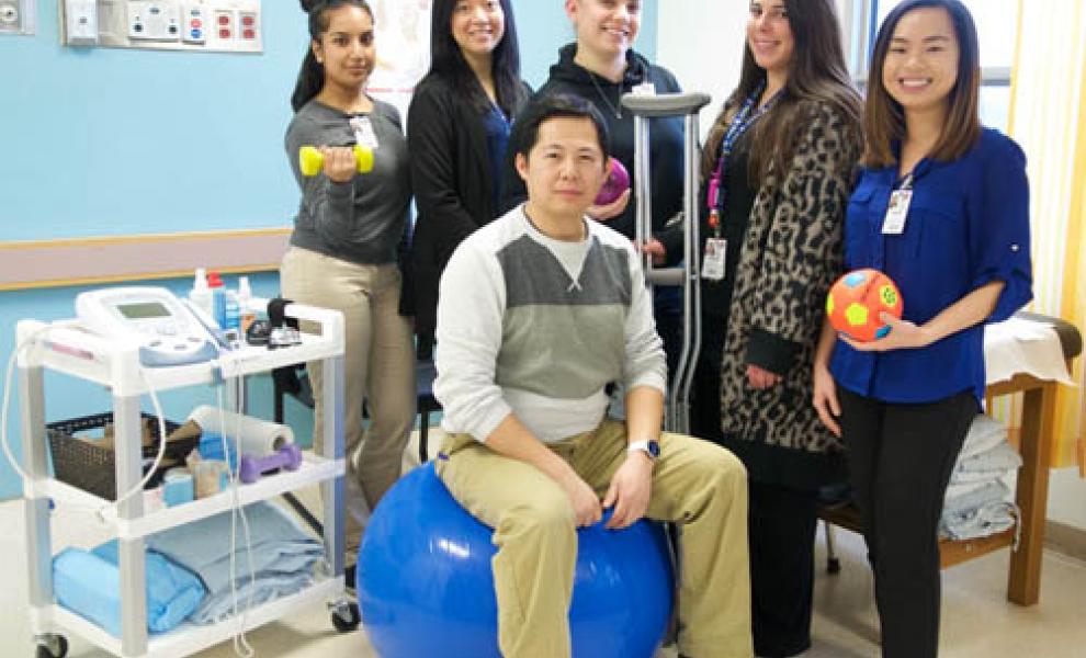 the physio therapy team of 6 people pose together smiling. One team member holds crutches, one holds a weight, one holds a ball, and another sits on a large yoga ball.
