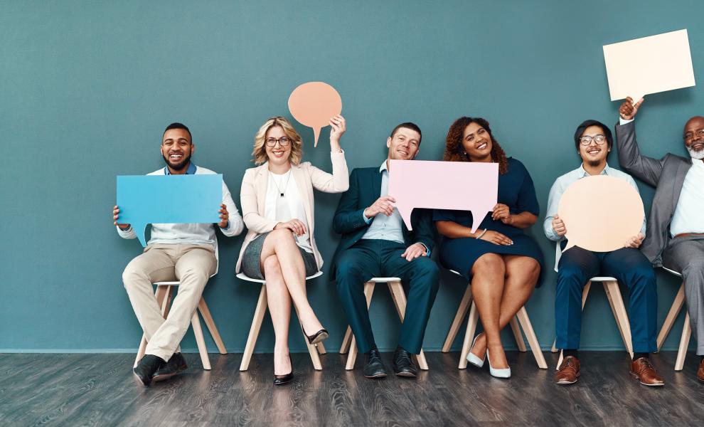 A diverse group of people sit in chairs smiling and holding up pieces of paper in shapes like squares or speech bubbles
