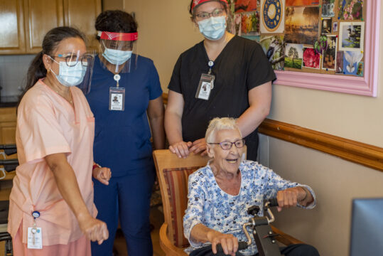 3 health care workers standing behind a senior who is operating an exercise bike.